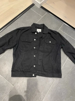 Svart jeansjacka från H&M - Stilren svart jeansjacka från H&M med klassisk krage, silverfärgade knappar och två bröstfickor med lock. Jackan har raka ärmar och en avslappnad passform som passar perfekt till alla typer av outfits. 
