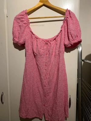 Rosa blommig kortklänning Miss Milla - Supersöt rosa kortklänning från Miss Milla med små vita blommor, puffärmar och knappar framtill. Klänningen har en smickrande v-ringning och resår vid axlarna för en bekväm passform. Perfekt för varma dagar!