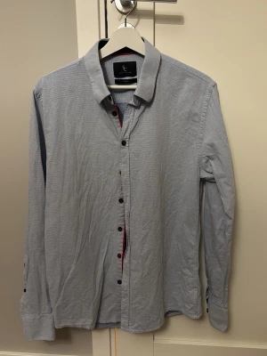 Blå skjorta från Savvy Citizen - Säljer en stilren blå skjorta från Savvy Citizen i storlek Large. Skjortan har ett diskret rutigt mönster, klassisk krage och svarta knappar. Materialet känns som bomull och den har långärmad passform. Perfekt för dig som vill ha en enkel men snygg look.