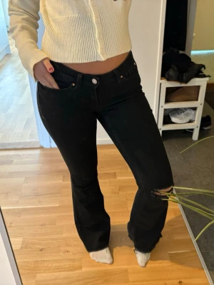 Lågmidjade Svarta bootcut jeans med slitning - Säljer ett par svarta bootcut jeans med slitning vid knät. Jeansen har hög midja, klassisk femficksmodell och är i stretchigt denimtyg. Snygg passform som framhäver benen och ger en trendig look.