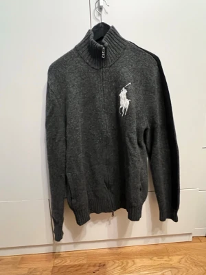 Grå stickad kofta Polo Ralph Lauren - SITTTER SOM EN L/M I STORLEKEN. Säljer en mörkgrå stickad kofta från Polo Ralph Lauren med stor vit logga på bröstet och 'GERMANY' broderat på ryggen. Tjock ribbad polokrage, hel dragkedja och patch med tysk flagga på ärmen. Perfekt för dig som gillar sportig och unik stil. 