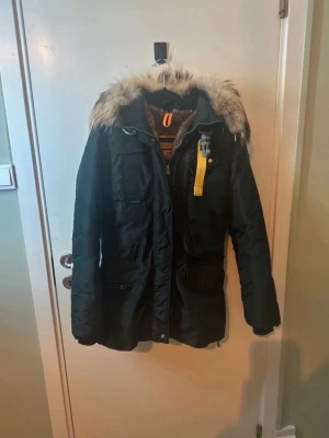 Svart parkas med päls från Parajumpers - Snygg svart parkas från Parajumpers med avtagbar fuskpäls på huvan och ikonisk gul detalj vid halsen. Jackan har flera fickor, dragkedja och knappar framtill samt ribbade muddar. Perfekt för kalla dagar och riktigt varm.storlek Xs men kan passa S kan eventuellt gå ner lite i pris vid snabb affär. 