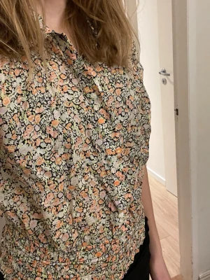 Blommig kortärmad blus med krage st 38 - Söt blommig blus från Oliver bonas med korta ärmar och klassisk krage. Mönstret går i pastelliga toner av orange, rosa, grön och svart på ljus botten. Blusen har knappar framtill och smockad midja för en snygg passform. Storlek 38