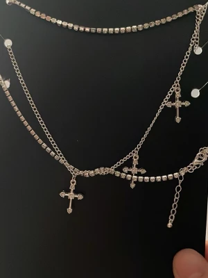 Silverfärgat halsband med korshängen - Trendigt halsband i silverfärg med flera lager och gnistrande stenar. Utsmyckat med små korshängen och kedjor i olika stilar. Perfekt accessoar för att lyfta din outfit med en edgy touch. Ordinarie pris 69kr.