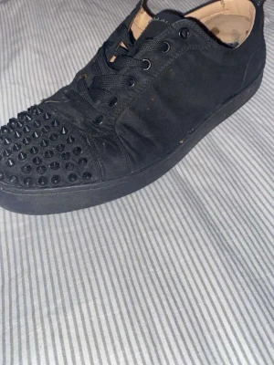 Svarta spikes-sneakers Louboutin - Säljer ett par svarta sneakers från Christian Louboutin med coola svarta nitar över tån. Skorna har klassisk låg profil, svarta skosnören och den ikoniska röda sulan. Materialet är tyg och detaljer i läder. Perfekt för dig som gillar stilrena skor nu till sommaren!!🤩.                            Priset är ej hugget i sten lägg bud!!!