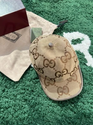Beige GG-mönstrad keps från Gucci - Säljer en beige keps från Gucci med det klassiska GG-mönstret i brunt. Kepsen har justerbart band i brunt skinn baktill och är tillverkad i ett slitstarkt tyg. Perfekt accessoar för dig som vill sticka ut med en lyxig touch.
