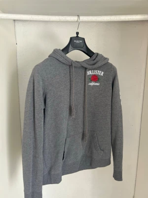 Grå hoodie från Hollister med tryck - Snygg grå hoodie från Hollister med broderad röd ros och texten 'California' på bröstet. Klassisk känguruficka framtill, justerbar huva med dragsko och ribbade muddar. Perfekt för en avslappnad och trendig look.