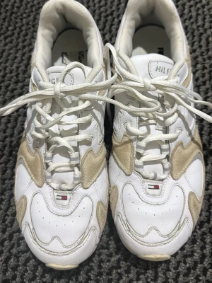 Vita sneakers från Tommy Hilfiger - Snygga vita sneakers från Tommy Hilfiger med beige mockadetaljer och klassisk logga på tån. Skorna har en chunky sula, vita snören och en sportig design. Perfekta för dig som gillar retrostil och streetwear.