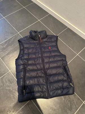 Mörkblå dunväst från Ralph Lauren Storlek S - Snygg mörkblå dunväst från Ralph Lauren med hög krage och dragkedja framtill. Västen har två fickor med dragkedja och den klassiska röda loggan broderad på bröstet. Perfekt för lager på lager och riktigt stilren look. Västen har inga skador är liknade. Storlek S