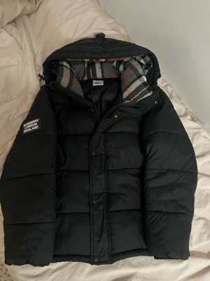 Burberry jacka - Svart pufferjacka från Burberry med klassisk logga på ärmen och avtagbar huva. Jackan har rutigt innerfoder i grått, rött och vitt samt dragkedja och knappar framtill. Perfekt för kalla dagar med sin quiltade design och varma känsla.