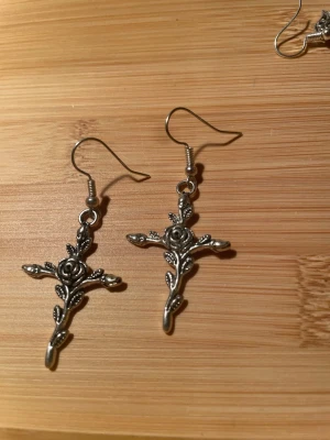 Silvriga gotiska örhängen med ros och kors - Handgjorda Unika silvriga örhängen i form av kors dekorerade med detaljerade rosor och blad. Hängande modell med krok, tillverkade i metall med snygg vintagekänsla. Perfekta för dig som gillar statement-smycken och coola detaljer. Ej nickelfria 