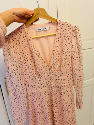 Ljusrosa blommig klänning från Adoore Paris  - Supersöt ljusrosa klänning från Adoore med små mörka blommor, v-ringning och knappar framtill. Klänningen har lång ärm och markerad midja för en smickrande siluett. Perfekt för vår och sommar! Nypris 1500kr