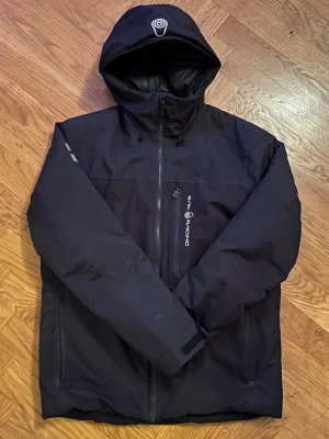 Sail Racing Jacka - Svart vindjacka från Sail Racing med huva och dragkedja framtill. Jackan har en bröstficka med dragkedja och logga på vänster sida. Tillverkad i slitstarkt syntetmaterial och har en enkel, sportig design som passar perfekt till kyliga dagar.