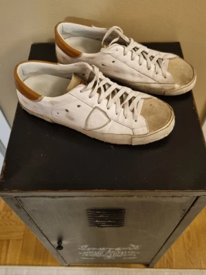 Vita Philippe Model sneakers med bruna detaljer - Snygga vita sneakers från Philippe Model med bruna detaljer vid hälen. Klassisk låg modell med vita skosnören och gummisula. Ovandelen är i läder och har diskreta sömmar och paneler på sidorna. Perfekta för en stilren och avslappnad look. Storlek 43 box finns även 