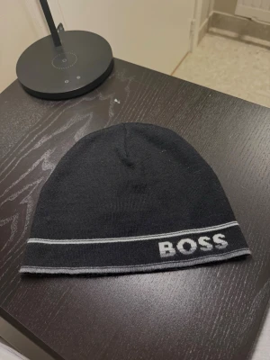 Svart mössa från BOSS med logga - Snygg svart mössa från BOSS med vit logga broderad framtill och tunna vita ränder längs kanten. Mössan är i mjukt stickat material och har en enkel, klassisk form som passar till det mesta. Perfekt accessoar för kalla dagar.