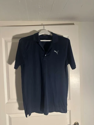 Marinblå pikétröja från Puma - Snygg marinblå pikétröja från Puma med klassisk krage och knappar framtill. Tröjan har korta ärmar och Puma-logga broderad på bröstet. Tillverkad i mjuk bomull för en bekväm känsla.
