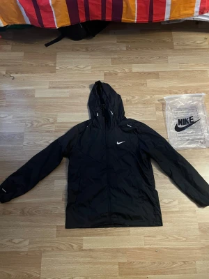 Svart vindjacka från Nike med huva - Snygg svart vindjacka från Nike med huva och dragkedja framtill. Jackan har diskret Nike-logga på bröstet och justerbara detaljer vid huvan. Perfekt för blåsiga dagar och enkel att matcha med olika outfits.
