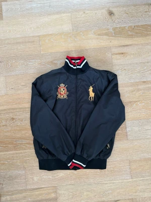 Svart vindjacka från Polo Ralph Lauren - Snygg svart vindjacka från Polo Ralph Lauren med broderad gul logga och emblem på bröstet. Jackan har ribbade muddar och krage med röd, vit och svart rand. Dragkedja framtill och fickor med dragkedja. Perfekt för en sportig och stilren look. Storlek L men passar också M