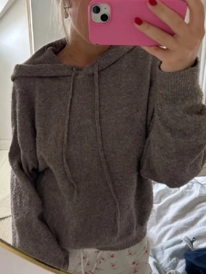 Brun hoodie med huva och dragsko - Mysig brun hoodie med huva och dragsko framtill. Tillverkad i ett mjukt, stickat material som ger en avslappnad och chill vibe. Långa ärmar med ribbade muddar och en lös passform som passar perfekt till vardagslooken.