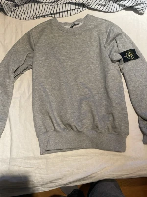 Grå sweatshirt från Stone Island - Snygg grå sweatshirt från Stone Island med den klassiska svarta patchen med gul och grön logga på vänster ärm. Tröjan har rund halsringning, ribbade muddar och är tillverkad i mjukt bomullsmaterial. Perfekt för en clean och avslappnad stil.