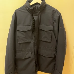 Mörkblå jacka från J.Lindeberg - Stilren mörkblå jacka från J.Lindeberg med fyra stora fickor framtill och hög krage. Jackan har dragkedja och knappar, samt diskret logga på ärmen. Perfekt för dig som gillar en klassisk look med praktiska detaljer.