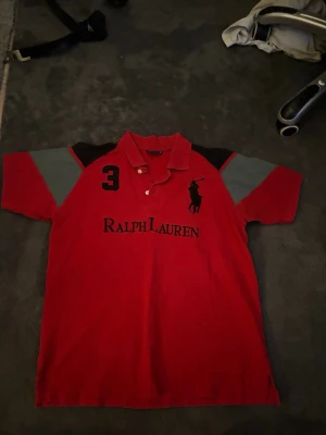 Röd Ralph Lauren pikétröja - Röd pikétröja från Ralph Lauren med svart logga och nummer 3 på bröstet. Tröjan har svarta och grå detaljer på axlarna samt klassisk krage och knappar framtill. Perfekt för en sportig och stilren look.