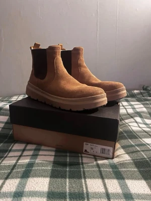 Bruna Chelsea boots från UGG - Säljer ett par snygga Chelsea boots från UGG i ljusbrunt mocka med elastiska svarta paneler på sidorna och grov sula. Skorna har dragflikar både fram och bak för enkel påtagning. Perfekta för dig som vill ha både stil och komfort.