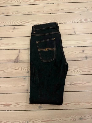 Mörkblå jeans från Nudie Jeans - Säljer ett par mörkblå jeans från Nudie Jeans med klassisk femficksmodell och kontrastsömmar. Jeansen har en slim passform och är tillverkade i kraftigt denimtyg av bomull. Snygg detalj med Nudie-loggan broderad på bakfickan.