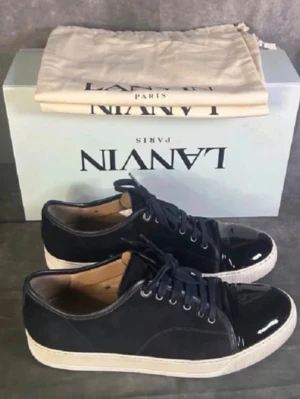 Svarta Lanvin sneakers med lackdetaljer - Snygga svarta sneakers från Lanvin med ovandel i mocka och glansiga lackdetaljer på tån. Skorna har vita gummisulor och svarta skosnören. Klassisk låg modell som passar till många olika stilar. Kommer med originalkartong och dustbag.