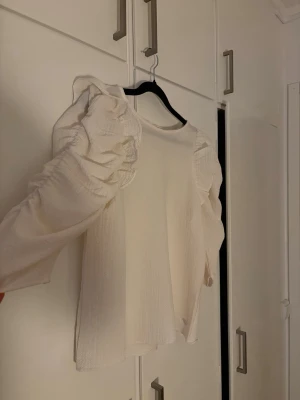 Vit blus med puffärmar och volanger - Trendig creme vit blus med långa puffärmar och volangdetaljer längs axlarna. Tillverkad i ett lätt, strukturerat tyg som ger en luftig känsla. Perfekt för dig som vill ha en statement-blus med unik siluett och snygga detaljer.