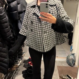 Kort kappa i svartvitt hundtandsmönster - Snygg kort kappa med klassiskt svartvitt hundtandsmönster och dubbelknäppning framtill. Kappan har bred krage och vida trekvartsärmar, vilket ger en trendig och avslappnad look. Perfekt för dig som vill sticka ut med en tidlös stil.
