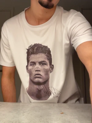 Vit t-shirt med porträtttryck - Säljer en vit t-shirt med stort porträtttryck på framsidan och en signatur under motivet. T-shirten har rund hals och korta ärmar, perfekt för dig som vill sticka ut med ett coolt print. Materialet känns mjukt och lätt, troligen bomull eller polyester.