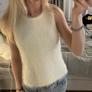 Vit fluffig ärmlös stickad topp - Supersöt vit stickad topp med fluffig känsla och ärmlös design. Perfekt till jeans eller kjol för en trendig look. Rund halsringning och mjukt material som känns skönt mot huden.