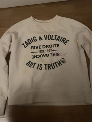 Vit sweatshirt från Zadig & Voltaire - Cool vit sweatshirt från Zadig & Voltaire med svart tryck på och texten 'ART IS TRUTH'. Tröjan har rund halsringning och långa ärmar. Materialet känns mjukt och bekvämt, perfekt för en avslappnad look. Den e kräm vit