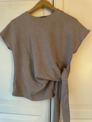 Beige topp med knytband - Säljer en stilren, kortärmad topp i en mjuk och behaglig taupe/mullvadsbrun färg. Toppen har en unik design med en dekorativ knut/knytdetalj i midjan som ger en mycket smickrande silhuett Helt ny
