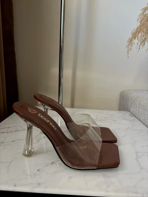Bruna klackade sandaler från Boohoo - Säljer ett par trendiga bruna sandaler från Boohoo med transparenta remmar och fyrkantig tå. Klacken är i klar plast och ger en modern look. Perfekt för dig som gillar stilrena och coola skor. Materialet är syntet och plast.