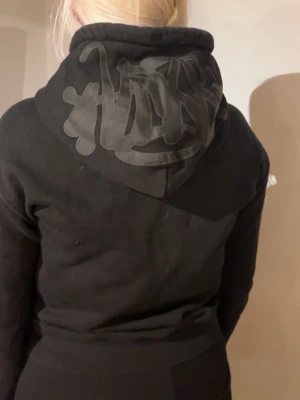 Svart märkes hoodie med tryck på huvan - Snygg svart hoodie med coolt svart tryck på huvan. Hoodien har en klassisk passform och är tillverkad i mjukt material som känns skönt mot huden. Perfekt för en avslappnad och trendig look.