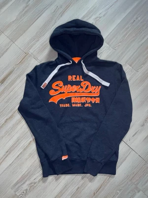 Mörkgrå Superdry hoodie med tryck - Snygg mörkgrå hoodie från Superdry med stort orange tryck på bröstet🤗 Längd: 71cm bredd: 57cm Storlek M kan även passa S🤩Skriv gärna vid snabbaffär samt funderingar. går jättegärna ner vid snabbaffär🙌🏻