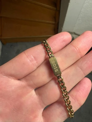 Guldfärgat armband från BOSS - Stilrent guldfärgat armband från BOSS med kraftig kedja och en rektangulär platta där BOSS är ingraverat. Armbandet har en klassisk och tidlös design i metall som passar perfekt till dig som gillar accessoarer med attityd. 60 cm 4mm