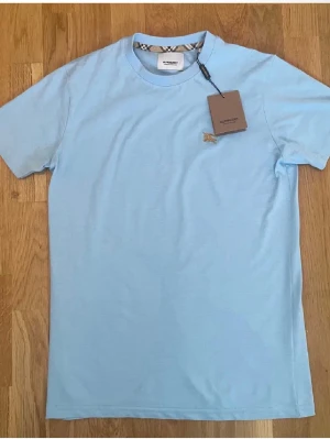 Ljusblå Burberry t-shirt i bomull - Snygg ljusblå t-shirt från Burberry med klassiskt rund halsringning och diskret Burberry-logga broderad på bröstet. T-shirten har korta ärmar och detaljer med Burberrys rutmönster på insidan av halsen. Tillverkad i mjuk bomull för en skön känsla.