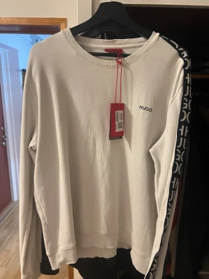 Vit sweatshirt från HUGO med logga - Snygg vit sweatshirt från HUGO med svart logga på bröstet och coola svarta band med vit HUGO-typografi längs ärmarna. Tröjan har rund halsringning och är tillverkad i mjukt material för en skön känsla. Perfekt för en trendig och avslappnad stil.