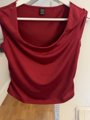Vinröd topp från SHEIN - Snygg vinröd topp från SHEIN med draperad urringning. Tillverkad i ett mjukt och glansigt syntetmaterial som ger en skön känsla. Perfekt att styla med jeans eller kjol för en trendig look.