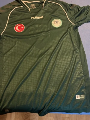 Grön Konyaspor matchtröja Hummel - Säljer en grön Konyaspor matchtröja från Hummel med vita detaljer och klubbmärke på bröstet. Tröjan har Turkiets flagga på ena sidan och är tillverkad i ett lätt, ventilerande material. Perfekt för fotbollsträning eller att bära på läktaren.