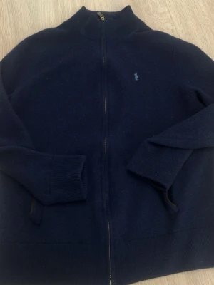 Mörkblå ziptröja från Polo Ralph Lauren - Mörkblå ziptröja från Polo Ralph Lauren med hög krage och diskret broderad logga på bröstet. Tröjan har långa ärmar, ribbade muddar och är tillverkad i mjukt stickat material. Perfekt för lager på lager och enkel att matcha. Storlek Xl men sitter som en M/L