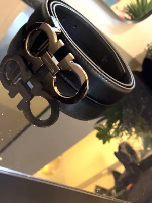 Ferragamo Gancini  - Stilrent svart skärp från Salvatore Ferragamo med ikoniskt dubbel-Gancini-spänne i metall. Skärpet är gjort i slätt skinn och har en elegant, tidlös design som passar till många olika outfits.