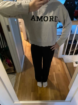 Grå/beige tröja med AMORE-tryck - Säljer en grå/beige stickad tröja med svart text 'AMORE' över bröstet. Tröjan har rund hals, långa ärmar och ribbade muddar vid ärmslut och nederkant. Är lite nopprig vid ryggens nedre mudd och har ett litet litet hål vid bröstet. Kontakta vid frågor