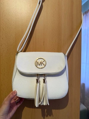 Vit axelväska från Michael Kors - Snygg vit axelväska från Michael Kors i strukturerat läder. Väskan har guldfärgade detaljer, två dekorativa tofsar och ett stort MK-emblem på locket. Justerbar axelrem och dragkedja framtill. Perfekt för dig som gillar stilrena accessoarer.