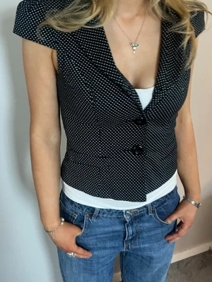 Svart prickig blus med knappar - Snygg svart blus med vita prickar och två stora knappar framtill. Modellen har korta ärmar och djup v-ringning. Figurnära passform som ger en chic siluett. Perfekt att styla med jeans eller kjol för en trendig look.