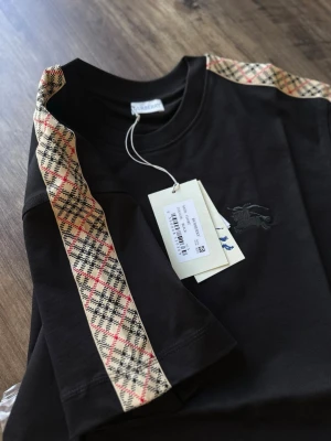 Svart Burberry t-shirt med mönstrade band - Svart t-shirt från Burberry med klassiska mönstrade band i beige, rött och svart längs axlarna. T-shirten har korta ärmar, rund hals och en diskret broderad logga på bröstet. Tillverkad i mjuk bomull för en skön känsla.