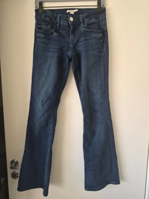 Blå bootcut jeans med låg midja - Snygga blå bootcut jeans med låg midja och klassiska fem fickor. Jeansen har en lätt tvättad look och vida ben från knät och ner. Perfekta för en trendig och avslappnad stil. Tillverkade i denim med stretch för extra komfort.